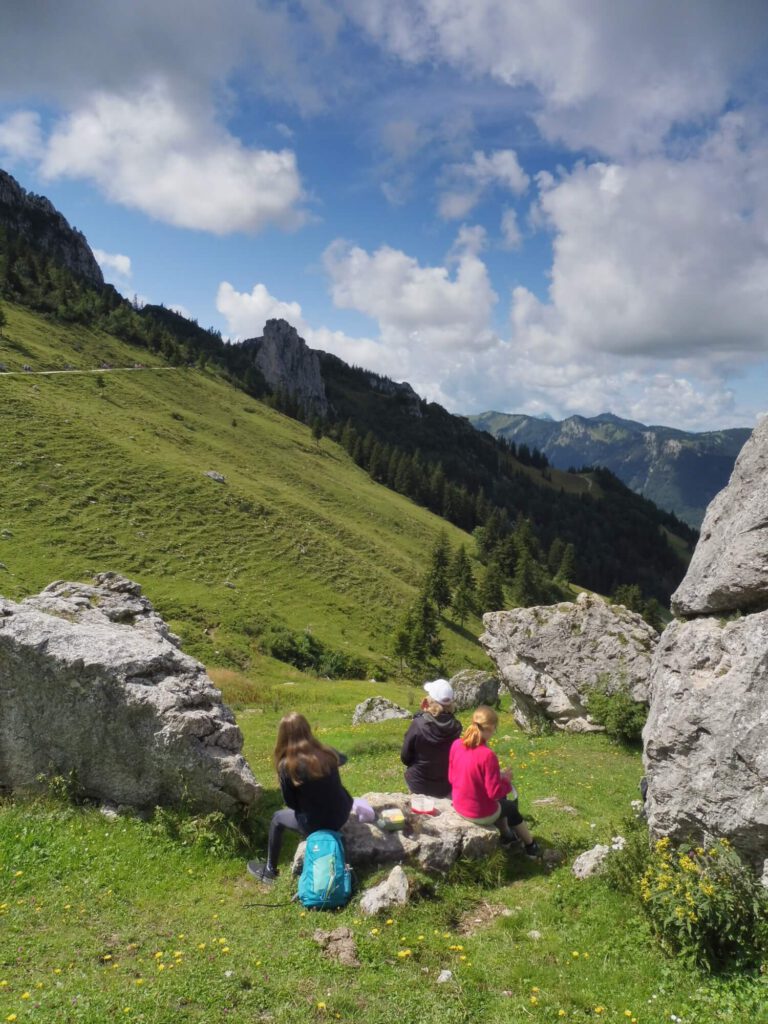 Foto dreier Menschen wie sie auf einer Bergwiese sitzend eine Pause machen. Ihre Blicken gehen zum Gipfel in der Ferne, den sie erreichen wollen.