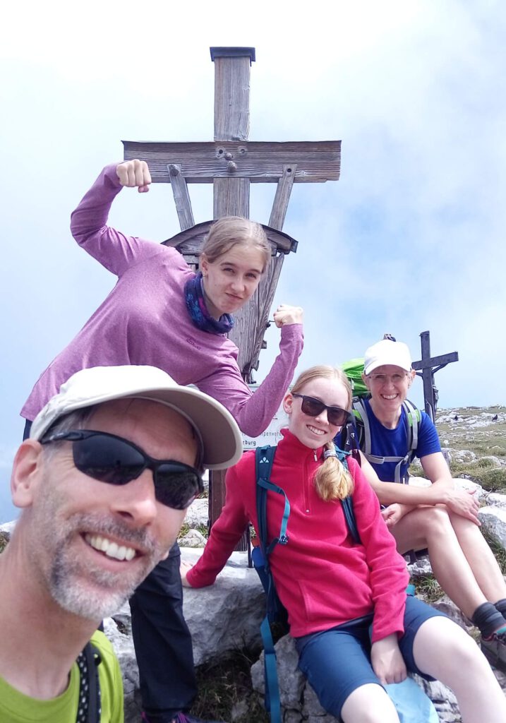 Foto unserer Familie vor einem Gipfelkreuz. Ausdruck von Triumph und Belohnung für die vorangegangenen Anstrengungen des Aufstiegs.