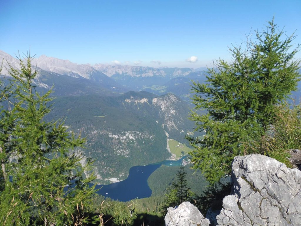 Foto einer Berglandschaft mit sanften, runden Bergen, einem Bergsee, Wäldern und grauen, kahlen spitzen Gipfeln.