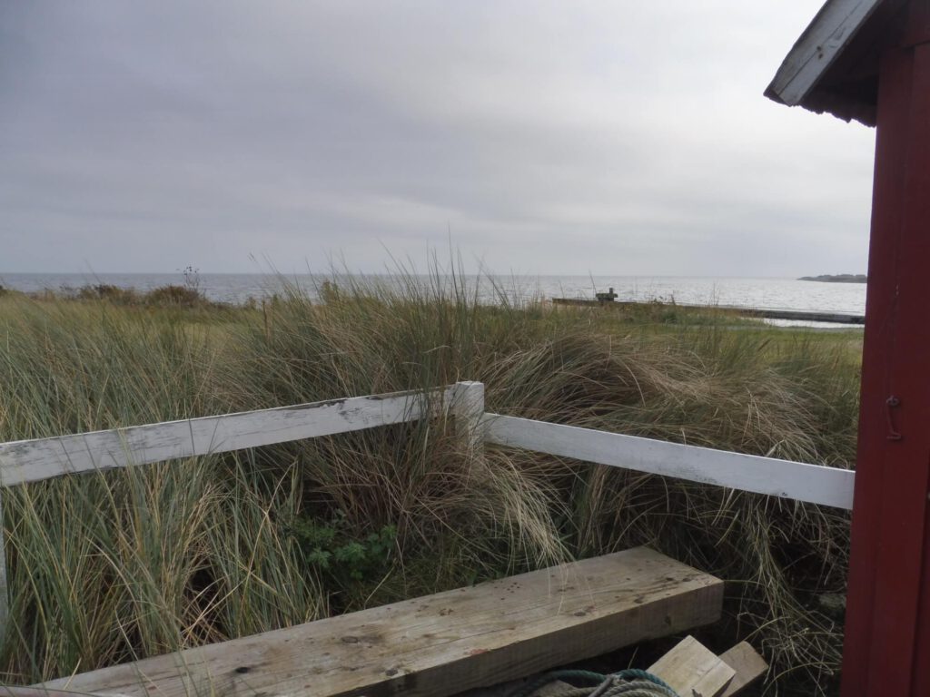 Foto Hausecke eines kleinen Hauses mit Veranda, dahinter das Meer. Kurzimpuls für Männer: Zitat von Udo Schroeter.