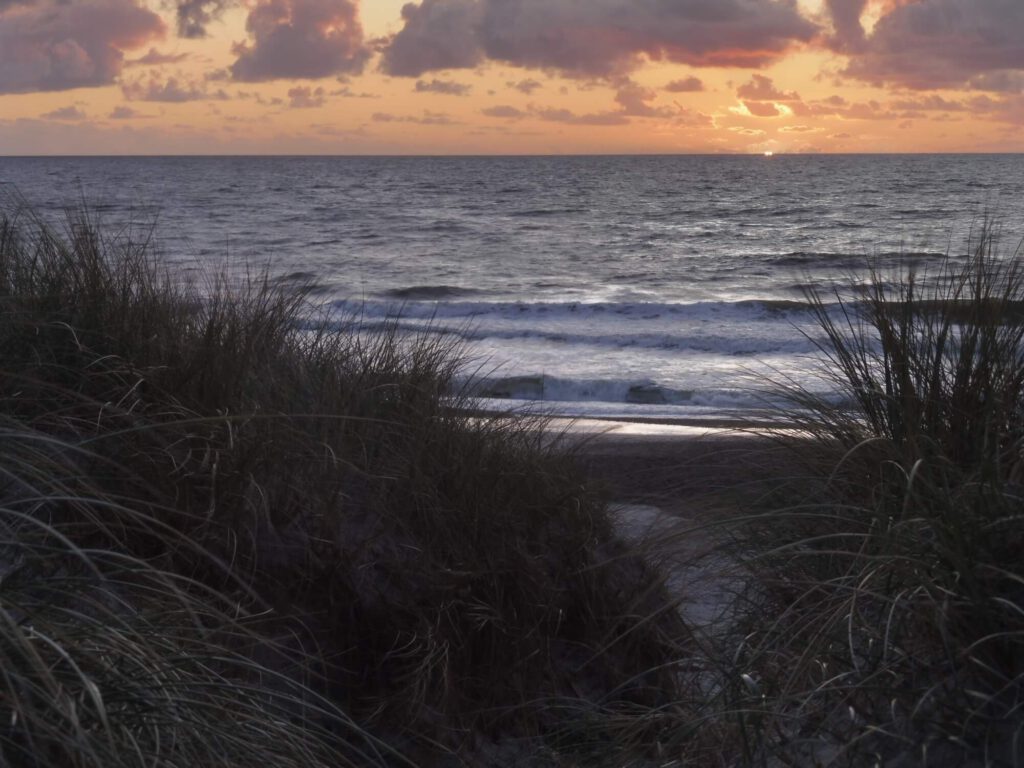 Foto eines Dünendurchgangs, mit Blick auf das Meer und die untergegangene Sonne.