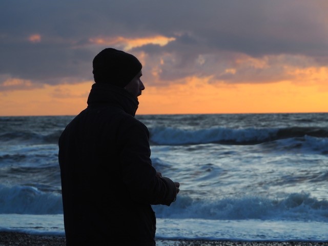 Bild eines Mannes, der auf das Meer hinaus blickt. Am Himmel Sonnenuntergangsstimmung, das Meer hat mächtig schäumende Wellen. Ausdruck von MEERwert für den Mann.