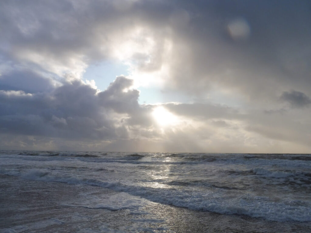 Die Sonne scheint auf das Meer herab durch ein Wolkenloch. Der Himmel ist blaugrau bedeckt, das Meer in ähnlichen Farben, mit weißem Schaum.