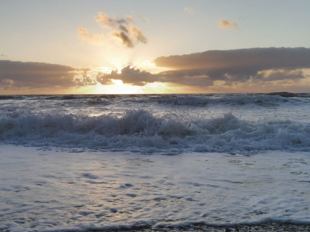 Bild eines beginnenden Sonnenunterganges am Meer. Im Vordergrund auslaufende spritzende Wellen mit Schaumkronen.