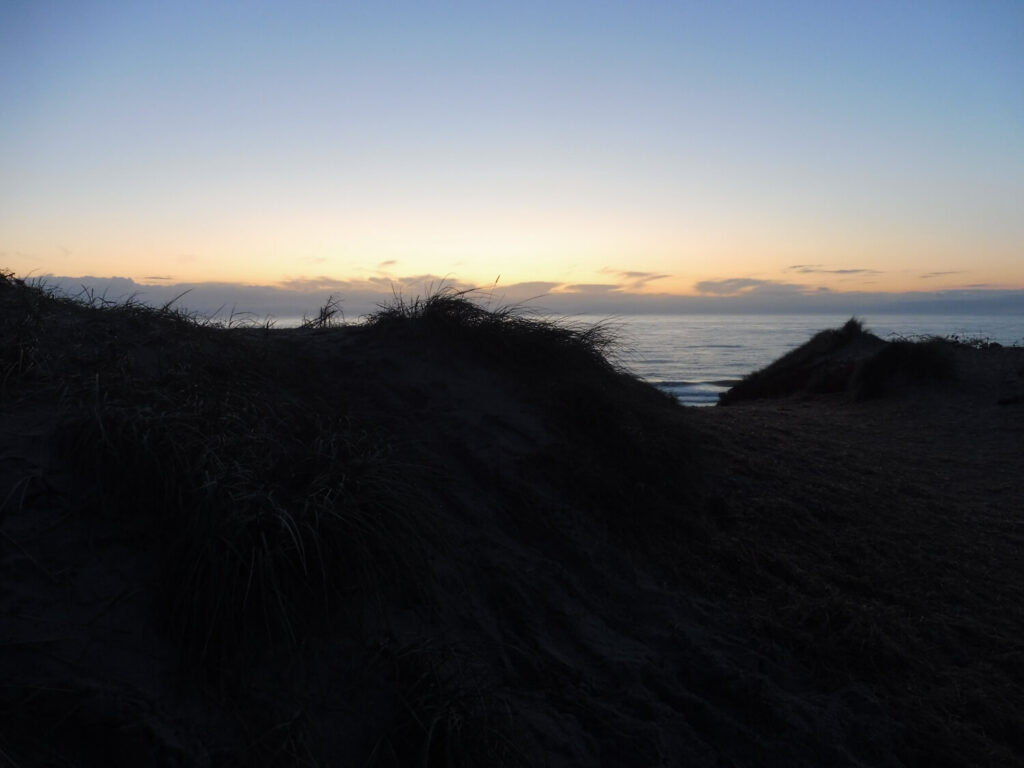 Bild einer Düne im Gegenlicht, dahinter das ruhige Meer.