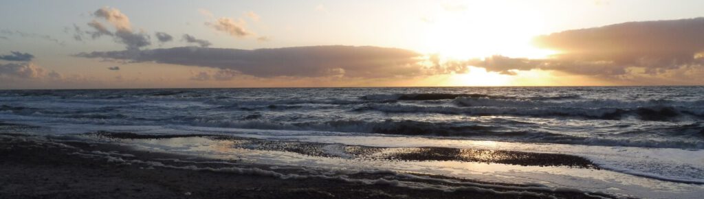 Bild vom Sonnenuntergang am Meer. Die Sonne bricht durch eine Wolke, in den Wellen spiegelt sie sich, das Meer leuchtet leicht orange. MEERwert durch Farben.