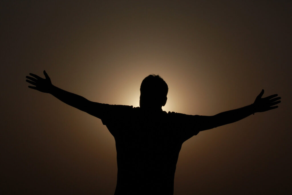 Foto eines Mannes, der im Gegenlicht beide Arme in den Himmel streckt. Symbolisch für "Ich habe eine Antwort auf die Frage "Wozu bin ich?"