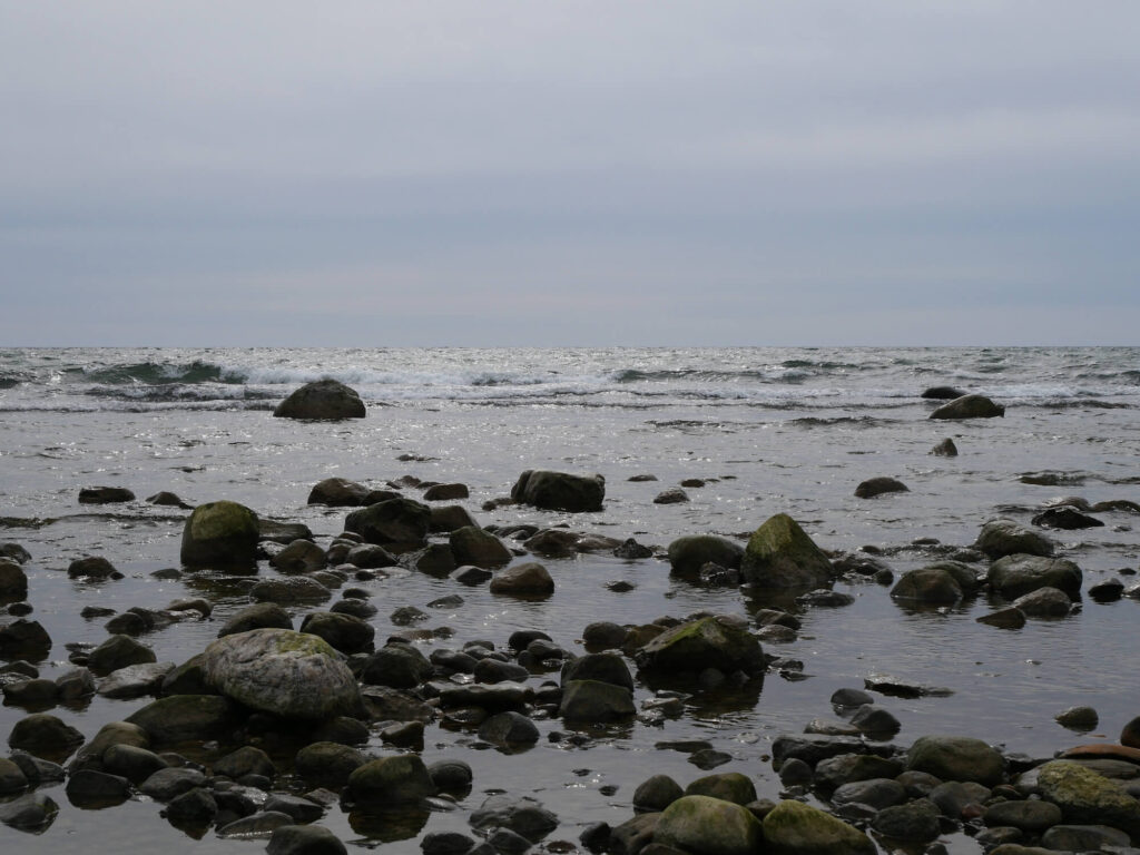 Mein Kraftort das Meer: Foto von Steinen im Strandbereich. Im Hintergrund brecken Wellen.